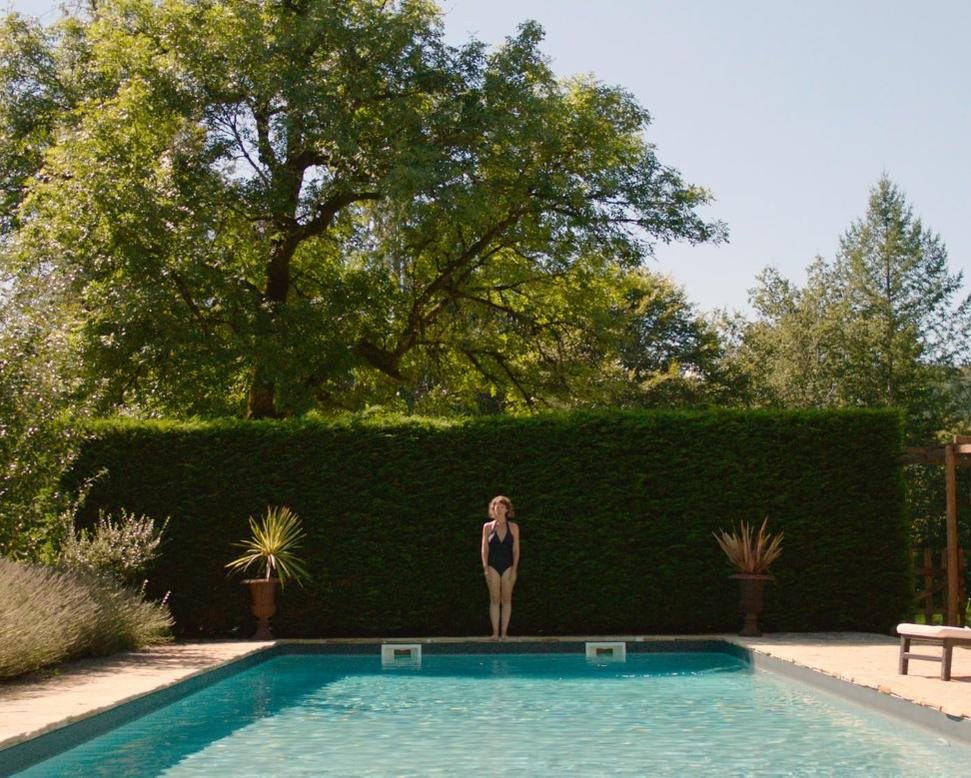 Eine Frau in einem Badeanzug steht am Pool vor einer Hecke.