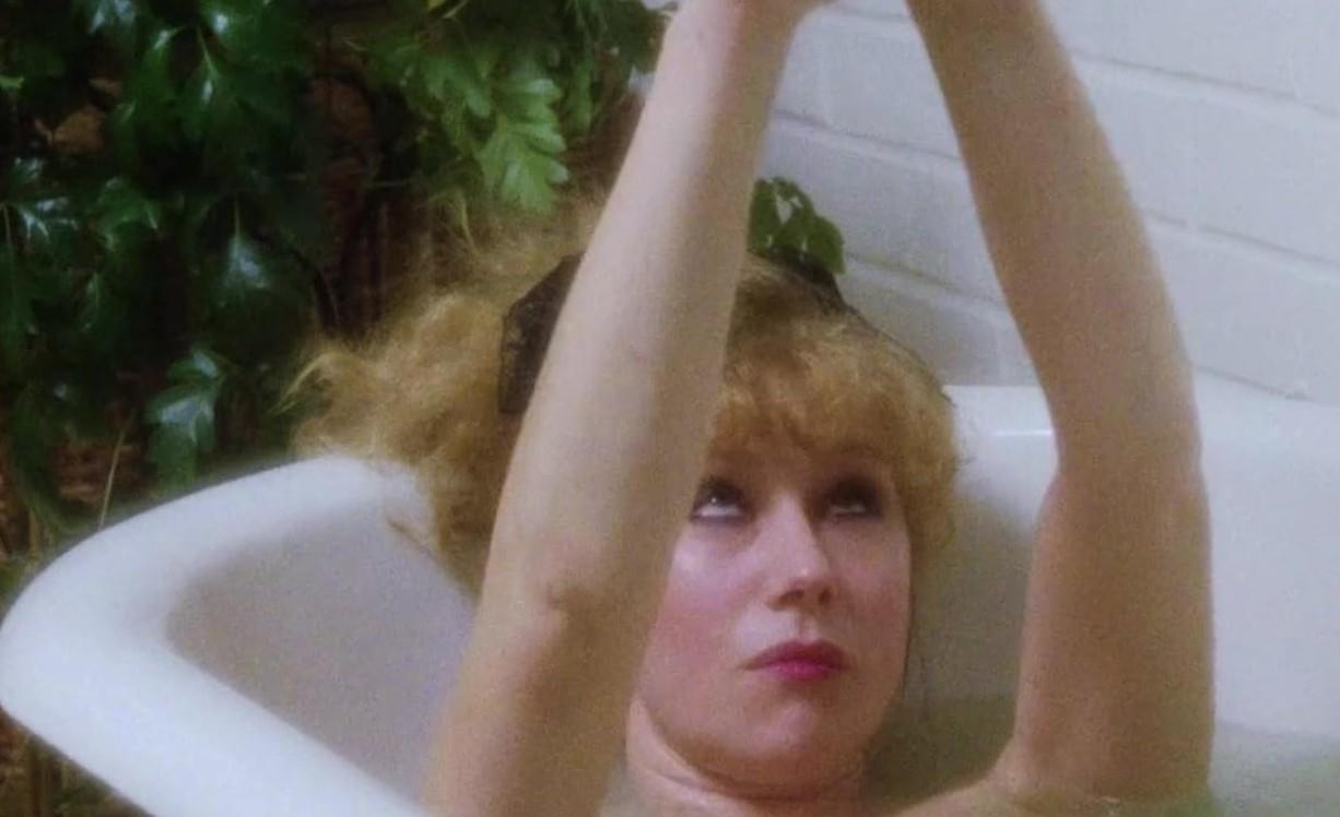 Blonde Frau mit erhobenen Armen in einer Badewanne, üppige grüne Pflanzen sichtbar.