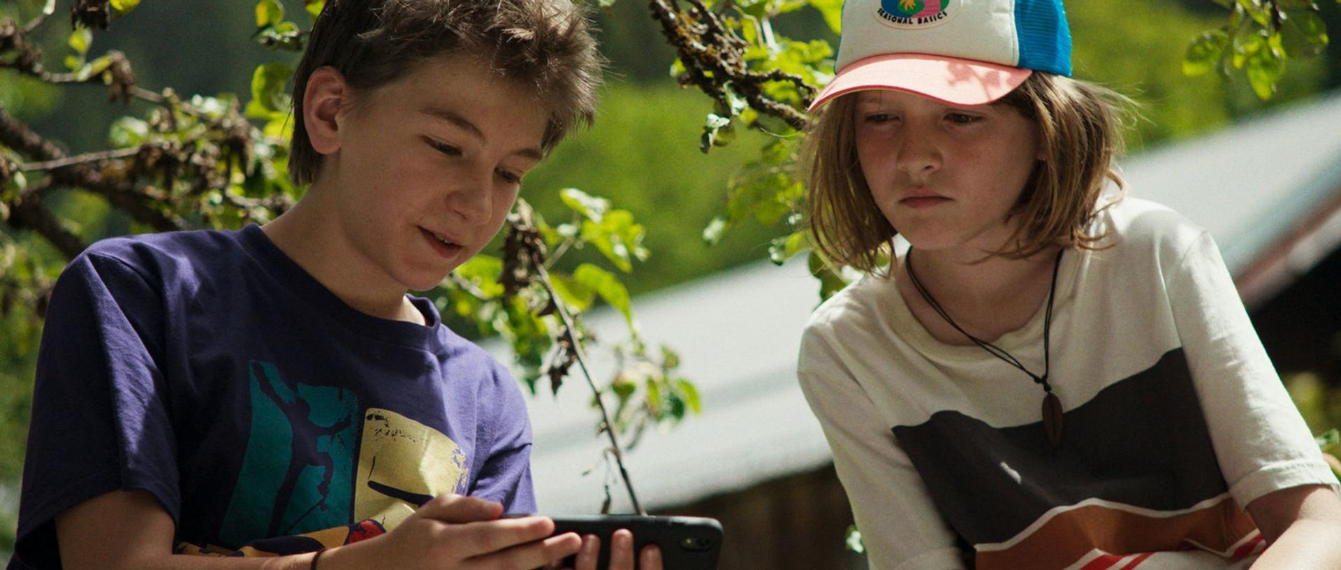 Zwei Jungen draußen, einer hält ein Smartphone, im Schatten von Bäumen.
