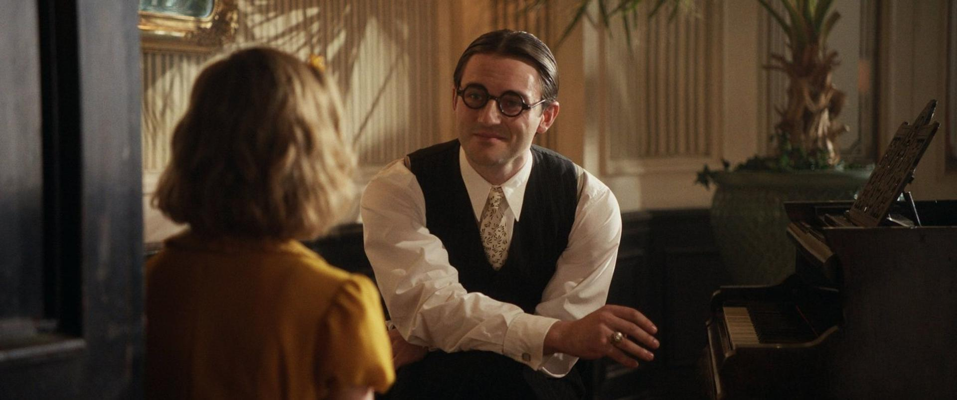 Ein Mann mit Brille und Weste spricht mit einer Frau in einem gelben Kleid neben einem Klavier.
