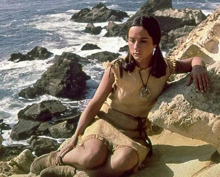 Junge Frau in Kleidung aus Naturmaterialien sitzt an felsiger Küste am Meer.