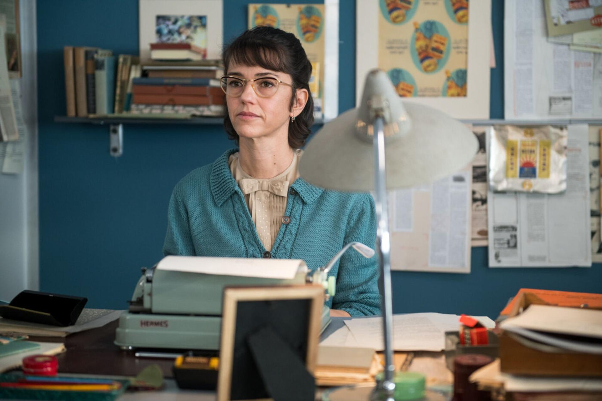 Eine Frau mit Brille sitzt an einem Schreibtisch mit einer Schreibmaschine und Büromaterial.