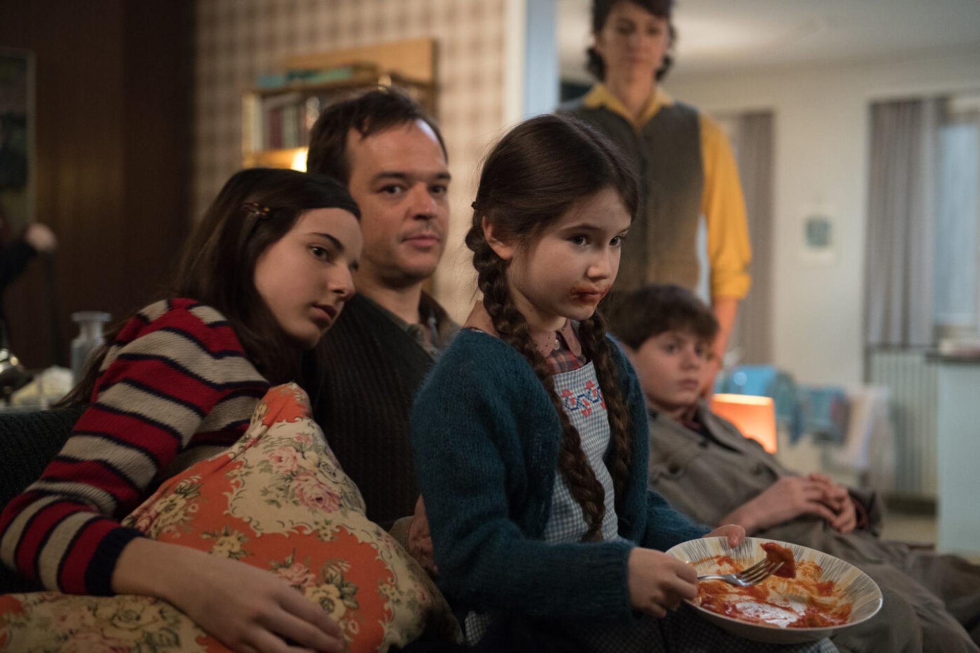 Eine Familie sitzt im Wohnzimmer. Ein Mädchen isst Pasta.
