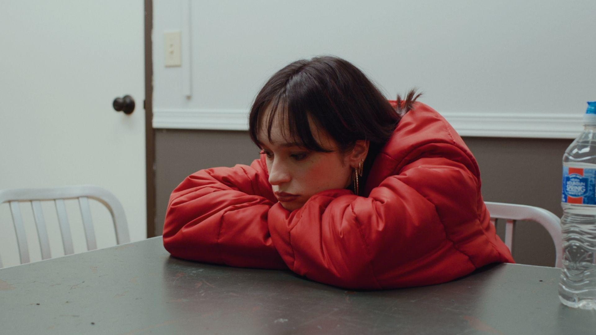 Junge Frau in roter Jacke mit Kopf auf Tisch, Wasserflasche rechts.