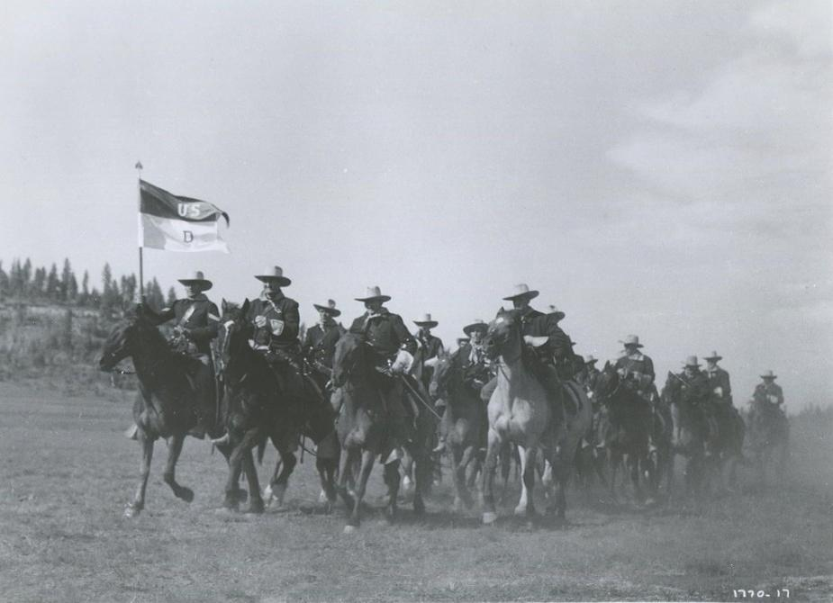 Eine Kavallerietruppe reitet über ein Feld, angeführt von einer Flagge. Soldaten tragen Hüte und Uniformen.