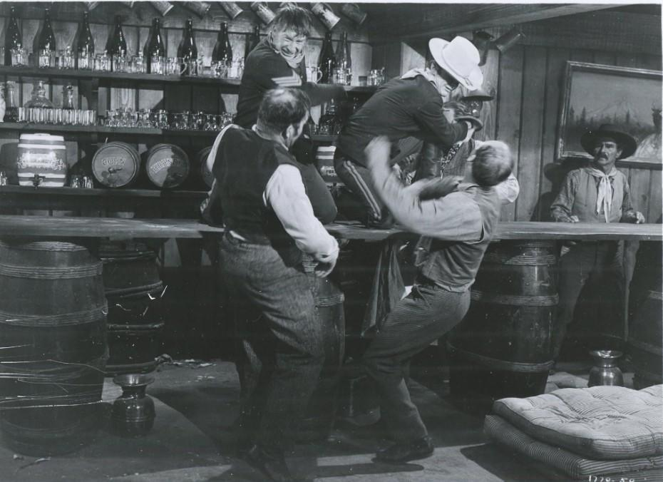 Fünf Männer in einem Saloon, einer wird über die Theke gehoben.