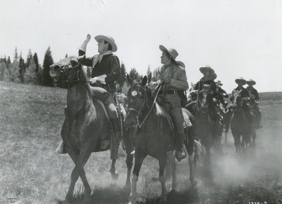 Eine Gruppe von Männern in Cowboyhüten reitet auf Pferden durch staubiges Gelände.