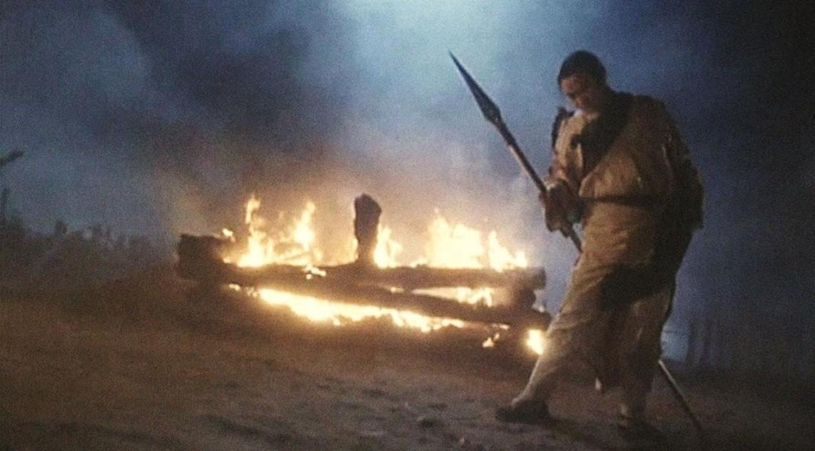 Mann mit Speer steht vor brennendem Holzstapel im Nebel.