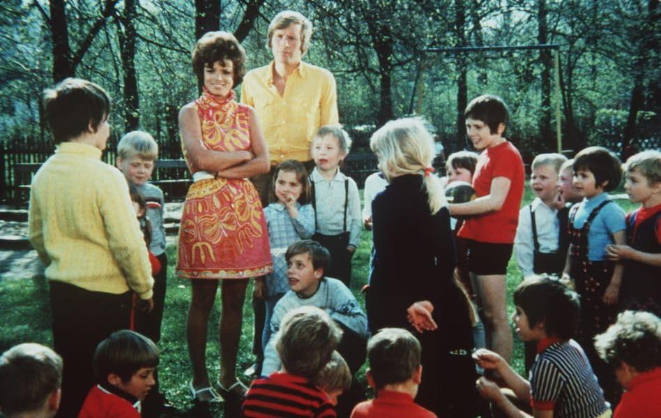 Eine Gruppe von Kindern und zwei Erwachsenen versammelt sich im Freien auf einer Grasfläche mit Bäumen.