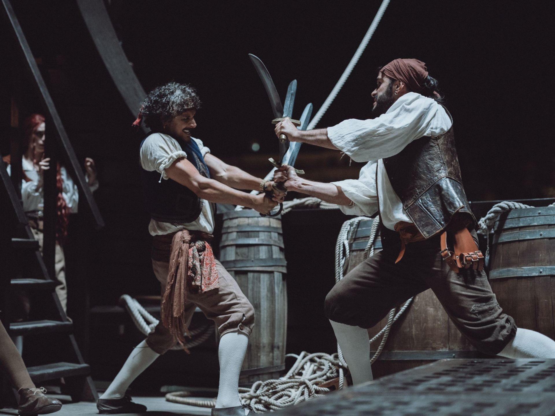 Zwei Männer in Piratenkostümen duellieren sich mit Schwertern auf einem Schiffsdeck mit Fässern.