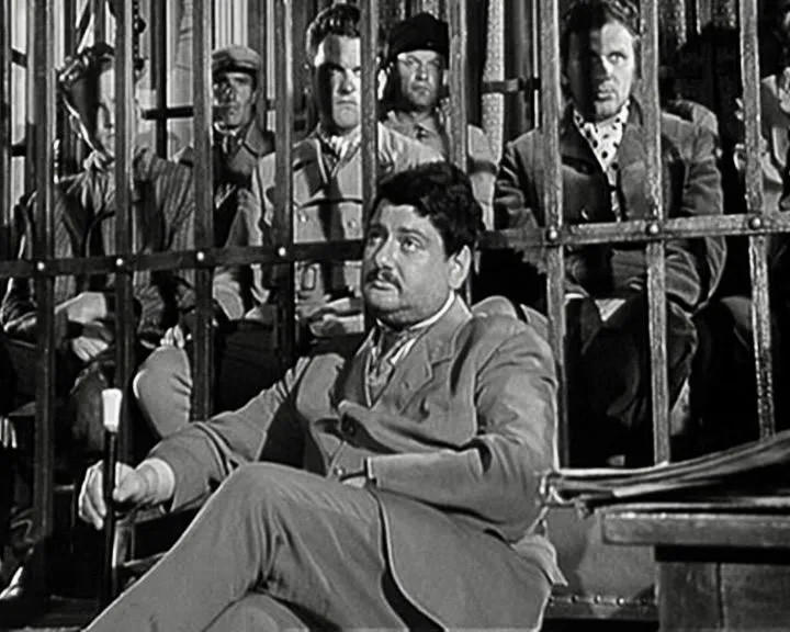 Männer in Anzügen sitzen hinter Gittern, einer stützt sich auf einen Stock.