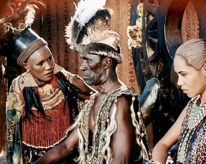 Drei Personen in traditioneller Kleidung mit Federschmuck und Perlen. Ein Mann in der Mitte, eine Frau links, eine Frau rechts.