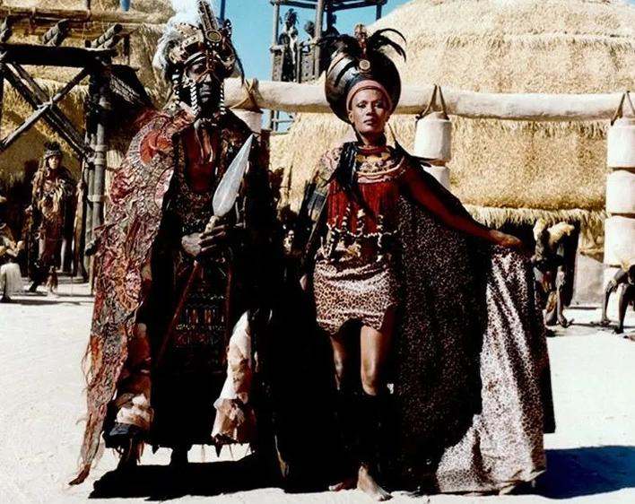 Zwei Personen in traditioneller Kleidung mit aufwendigen Kopfbedeckungen und Waffen.