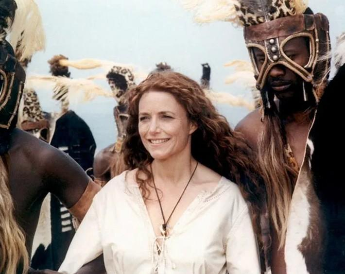 Eine Frau mit roten Haaren lächelt, umgeben von Männern in traditioneller Kleidung und Masken.