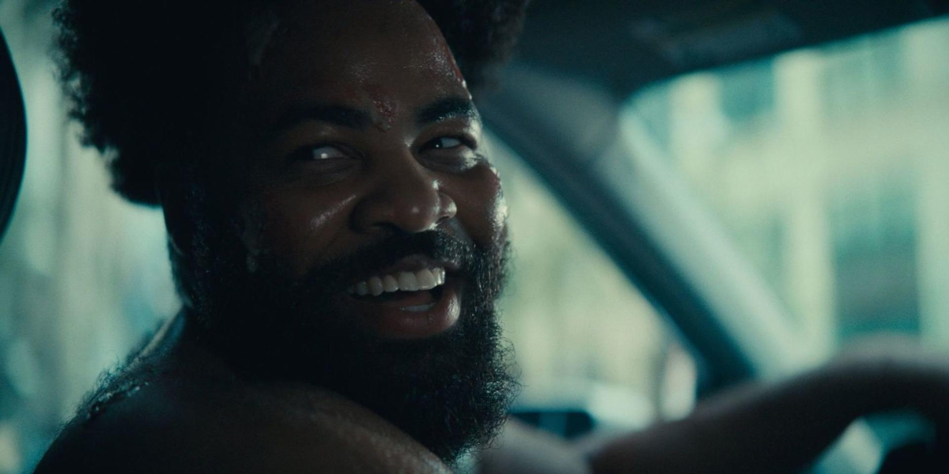 Ein Mann mit Bart und Afro lächelt in die Kamera, während er Auto fährt.