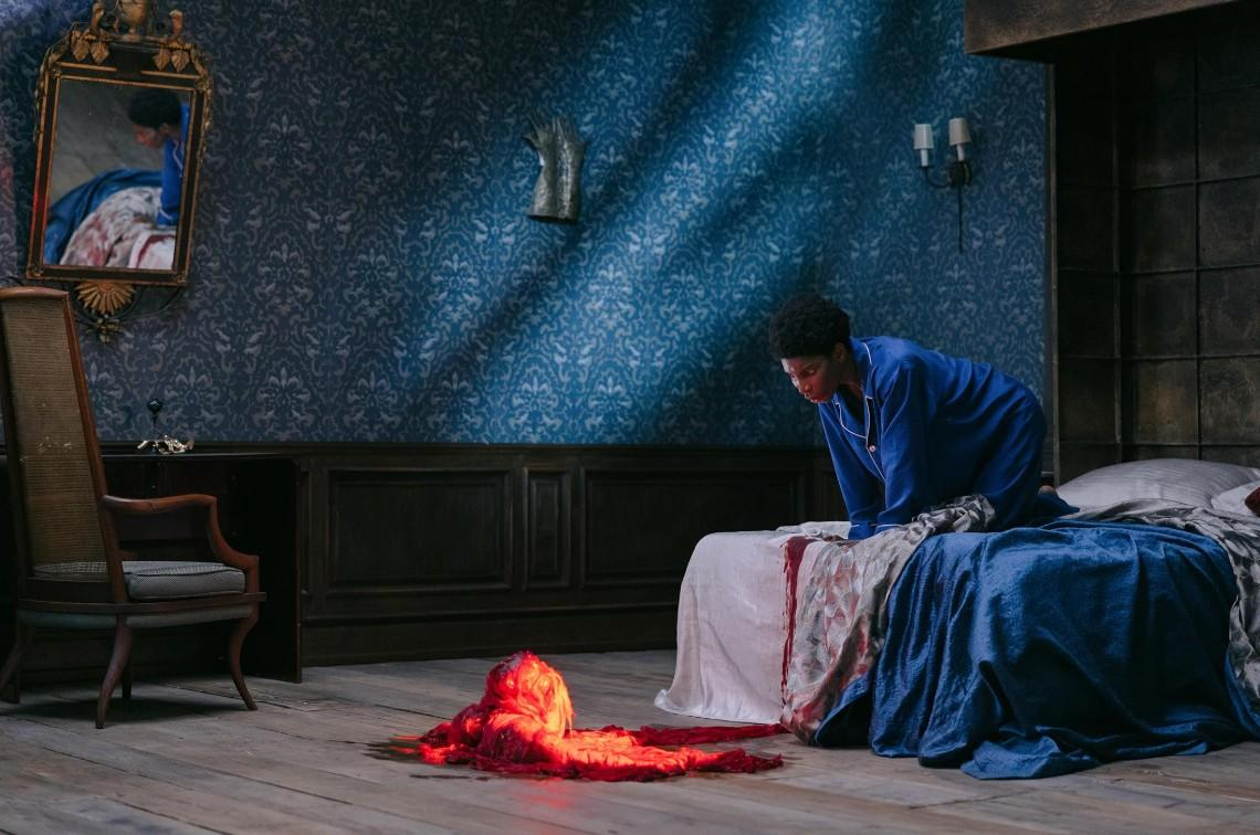 Eine Frau in blauer Kleidung kniet auf einem Bett neben einer roten, glühenden Masse auf dem Holzboden.