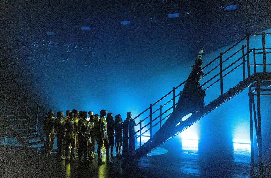 Eine Gruppe von Menschen steht auf einem dunklen Bühnenboden, während eine Person eine Treppe hinaufsteigt.