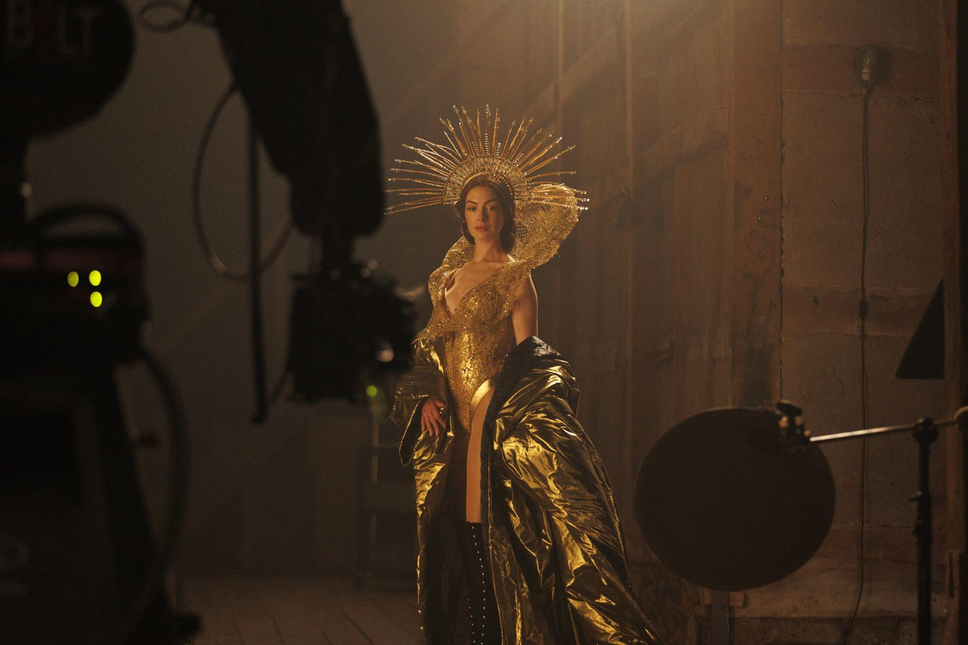 Eine Frau in einem goldenen Kleid mit einem strahlenden Kopfschmuck steht im Scheinwerferlicht.