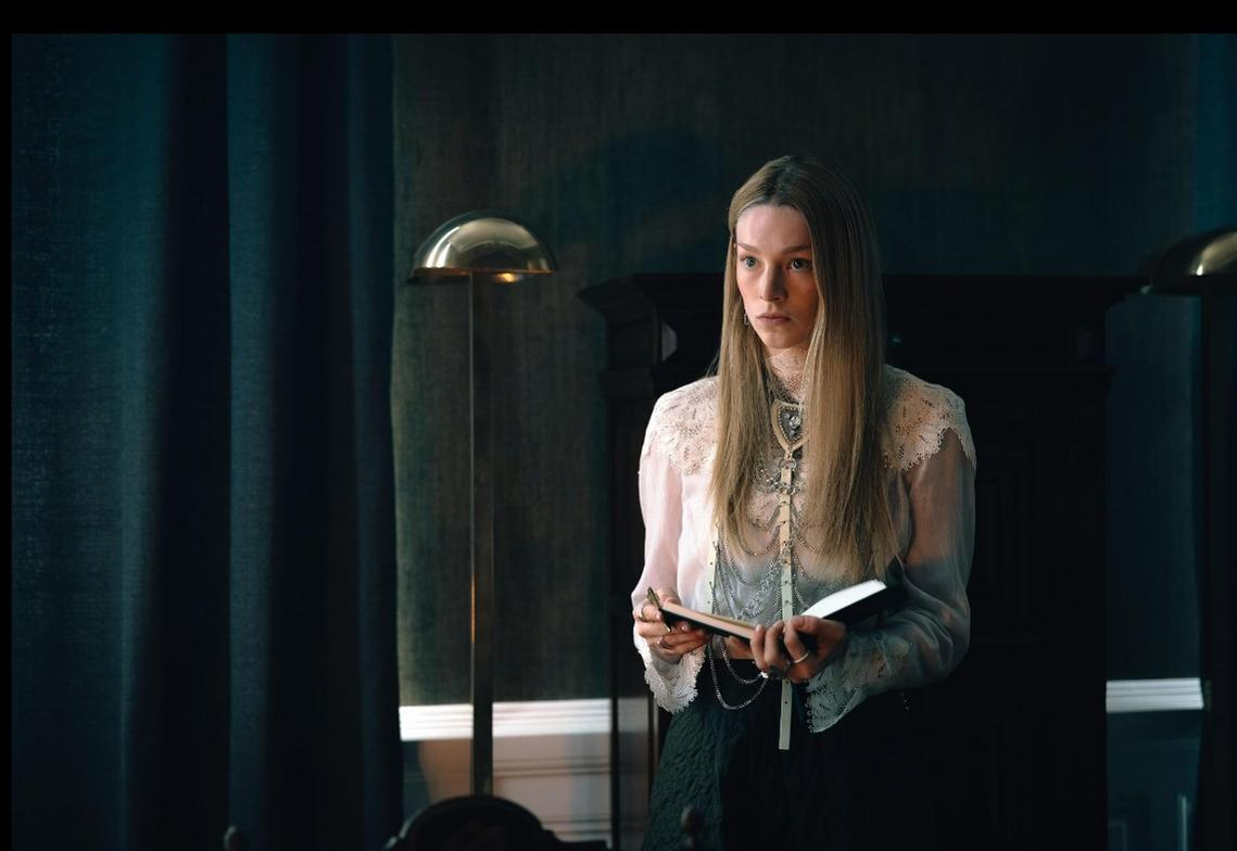 Eine Frau mit langen blonden Haaren hält ein Buch in einem schwach beleuchteten Raum mit dunklen Vorhängen.