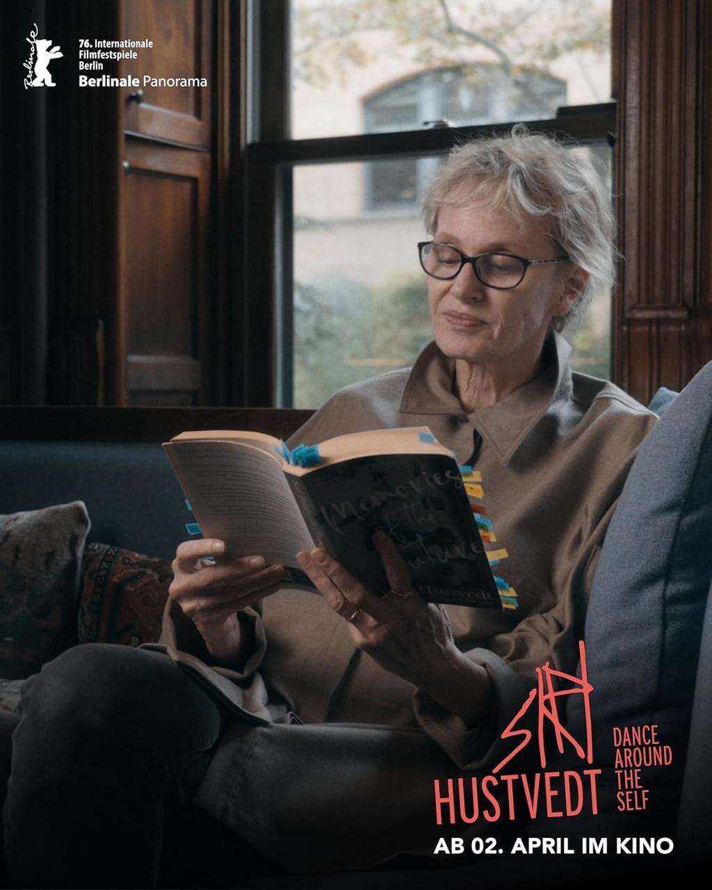 Eine ältere Frau mit Brille liest ein Buch auf einem Sofa. Im Hintergrund ist ein Fenster.