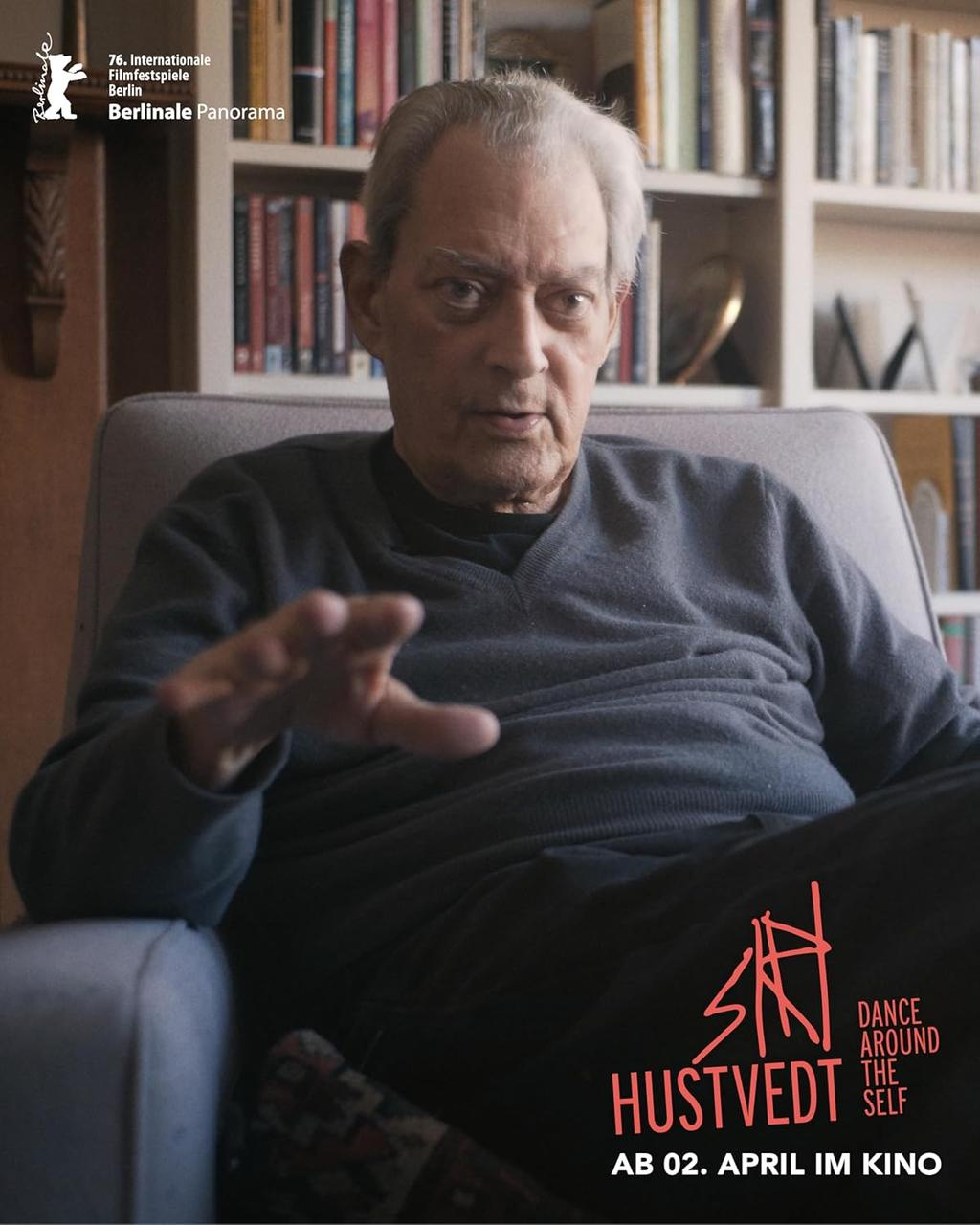 Älterer Mann im Sessel, spricht, mit Büchern im Hintergrund.