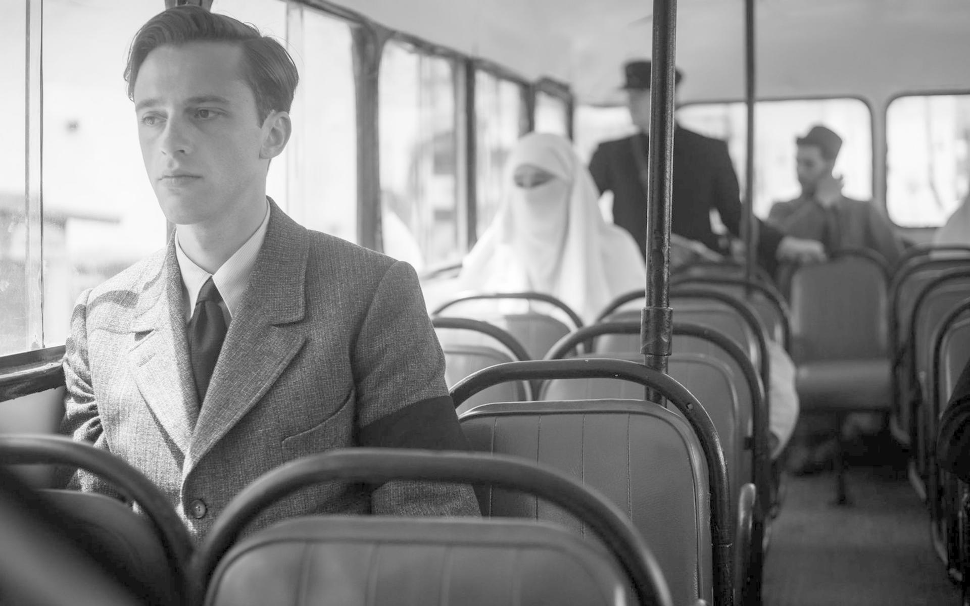 Ein Mann in einem Anzug sitzt in einem Bus. Andere Passagiere sind im Hintergrund zu sehen.