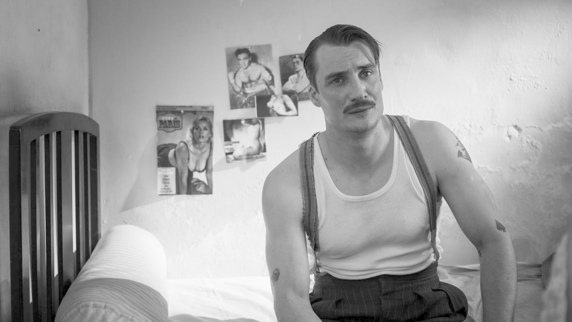 Ein Mann mit Schnurrbart sitzt auf einem Bett in einem Raum mit Pin-up-Postern an der Wand.