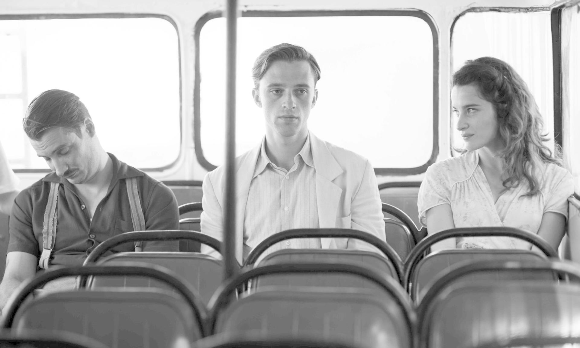 Drei Personen sitzen in einem Bus. Ein Mann schläft, ein Mann blickt nach vorne, eine Frau blickt zur Seite.