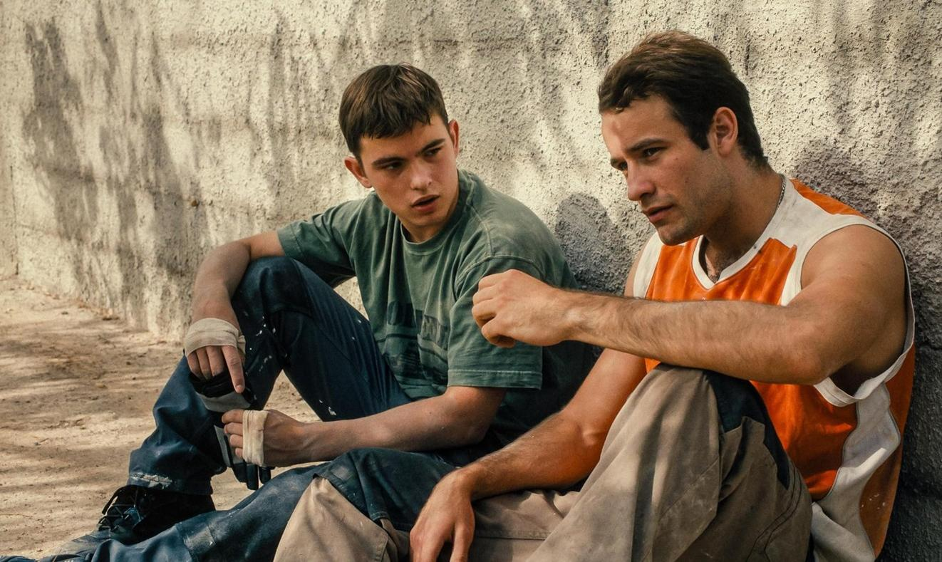 Zwei junge Männer sitzen an einer Wand. Einer trägt ein grünes Shirt, der andere ein orangefarbenes Tanktop.