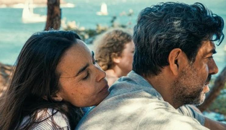 Eine Frau lehnt an einem Mann, im Hintergrund ein Kind und das Meer.