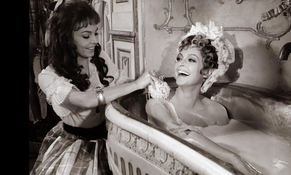 Zwei Frauen in Vintage-Kleidung teilen sich ein Bad. Eine wäscht die andere mit einem Schwamm.