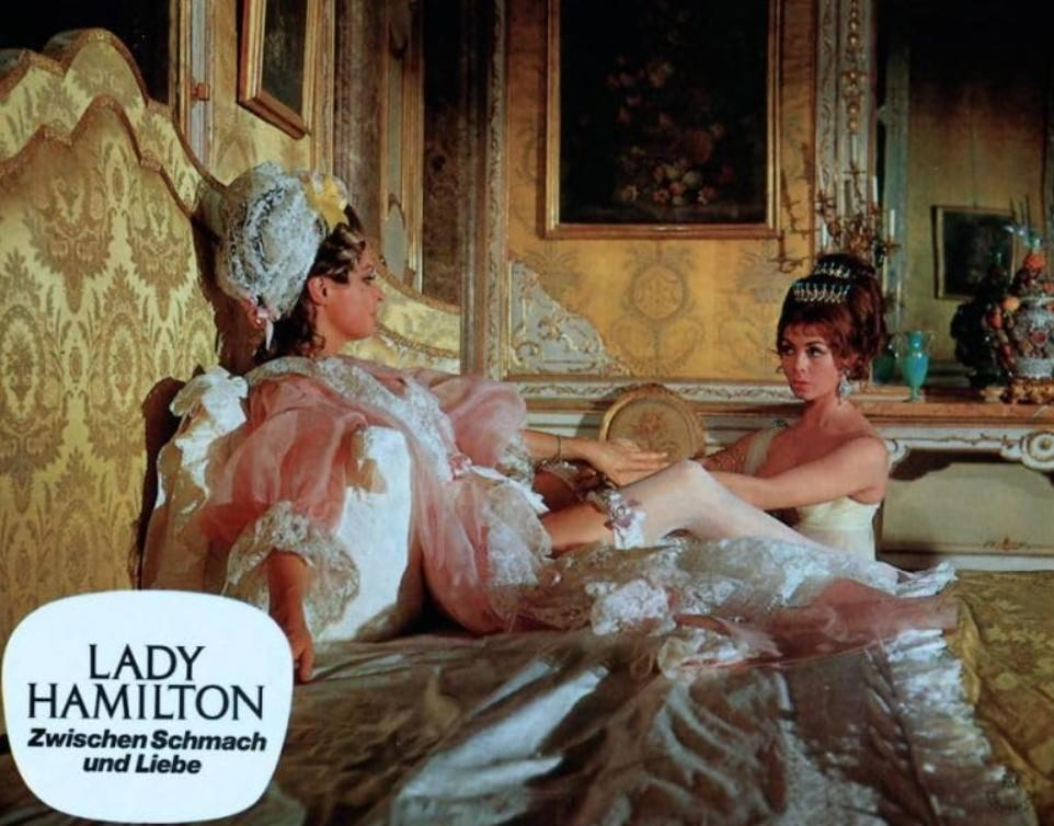 Zwei Frauen in historischen Kostümen liegen auf einem opulenten Bett in einem prunkvollen Raum.