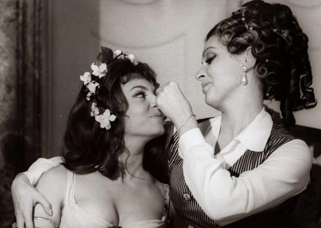 Zwei Frauen in historischer Kleidung küssen sich. Eine trägt Blumen im Haar.
