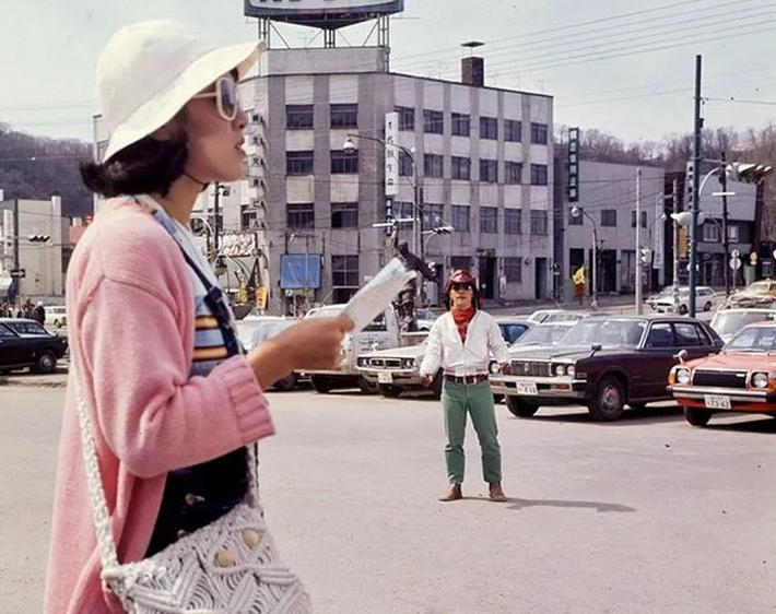 Eine Frau mit Hut hält eine Karte, ein Mann steht in der Nähe von Autos.
