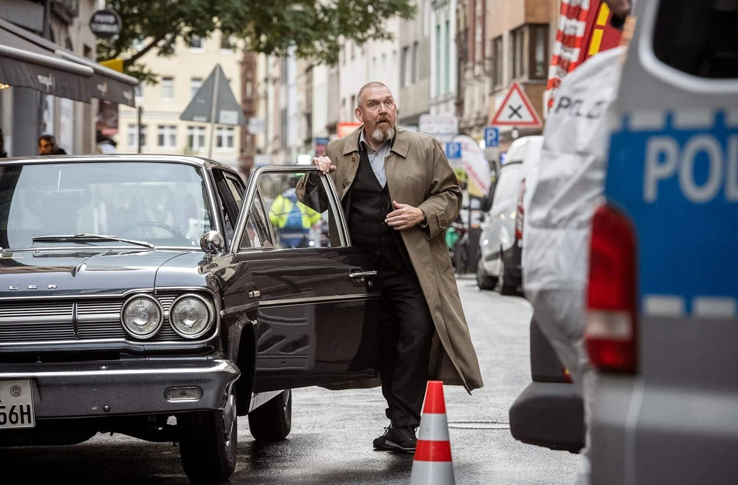 Ein Mann in einem beigen Trenchcoat steigt aus einem schwarzen Oldtimer.