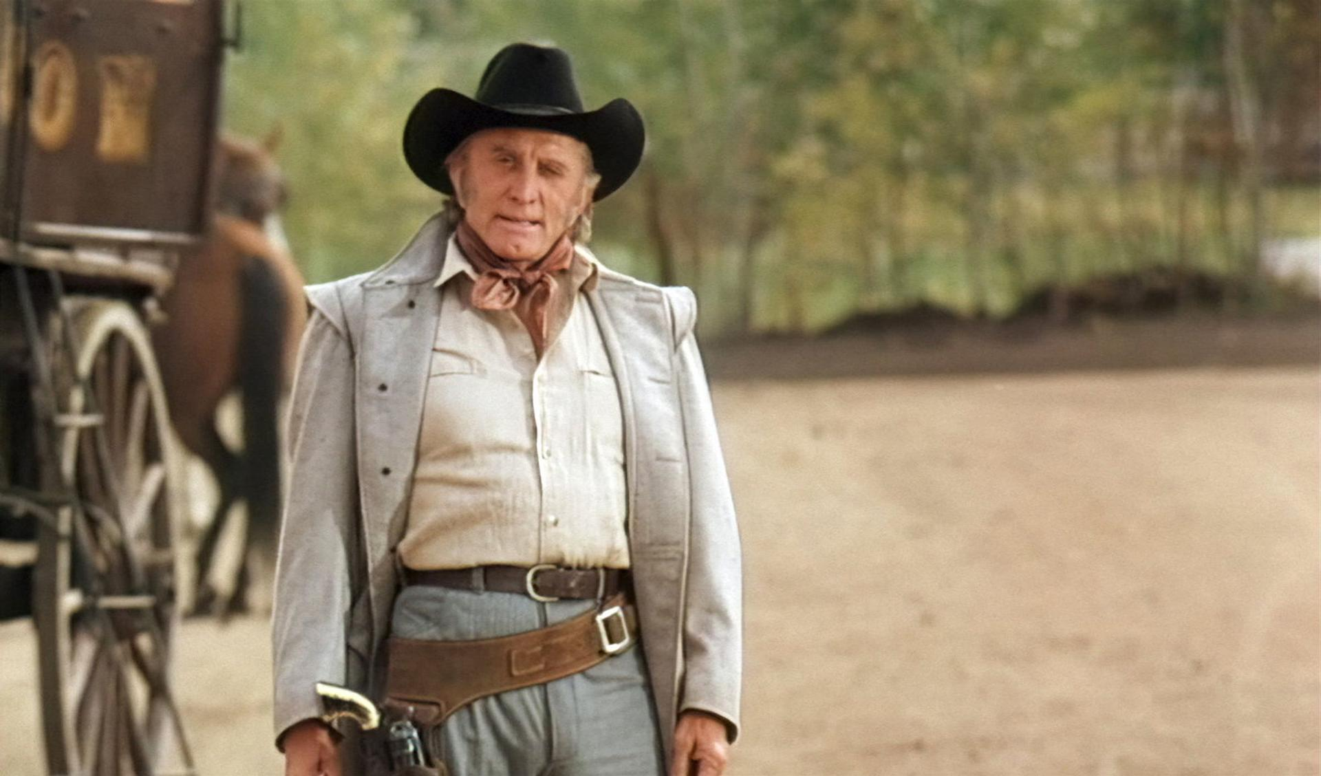 Ein Mann im Western-Outfit mit Hut und Revolver steht vor einem Wagen und Bäumen.