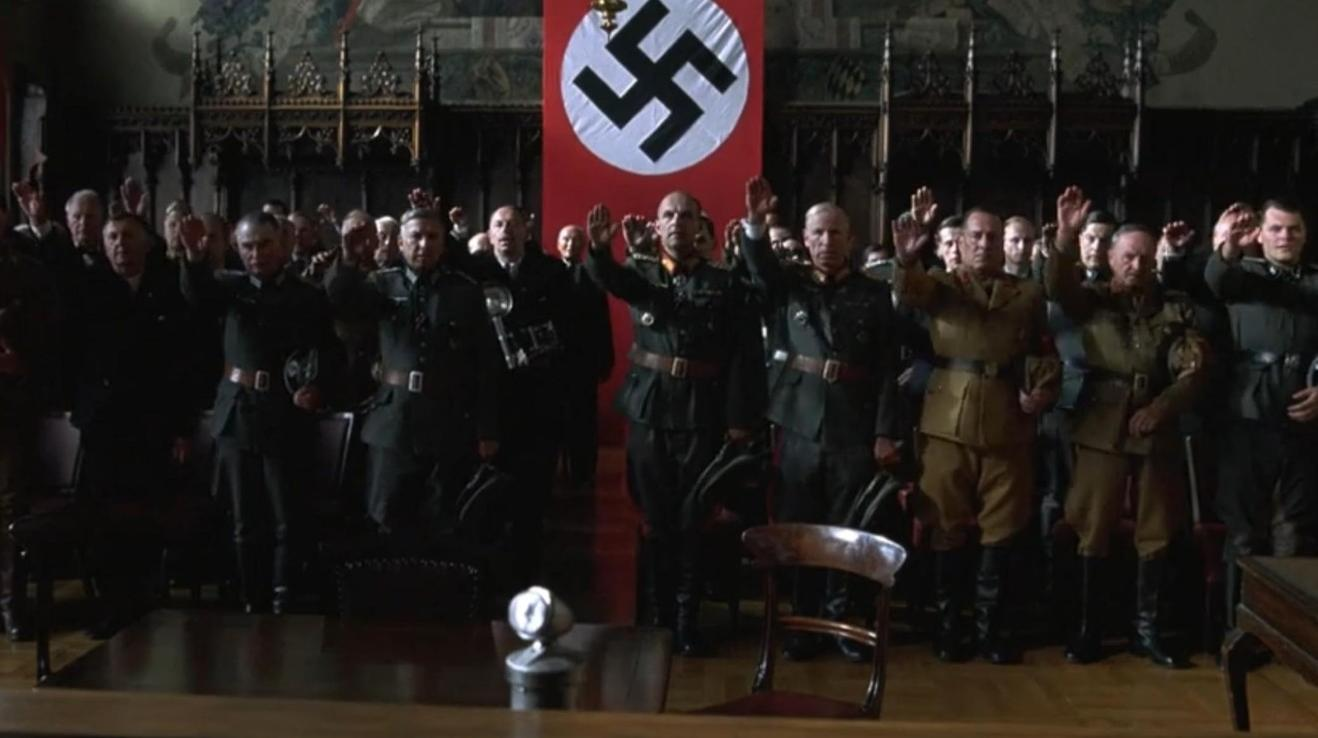 Männer in Uniformen salutieren vor einem Hakenkreuzbanner in einem großen Raum.