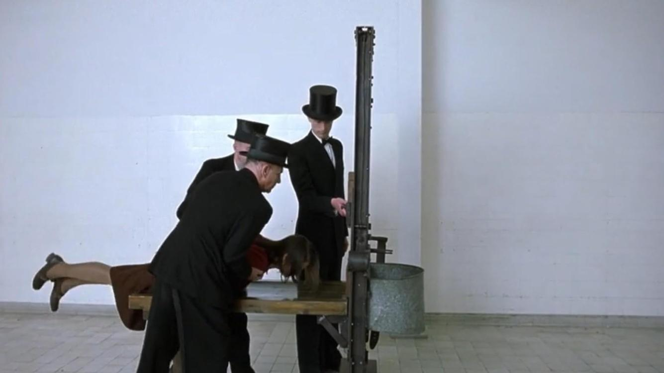 Drei Männer in Zylindern und Anzügen bedienen eine Guillotine an einer Frau, die auf einem Tisch liegt.