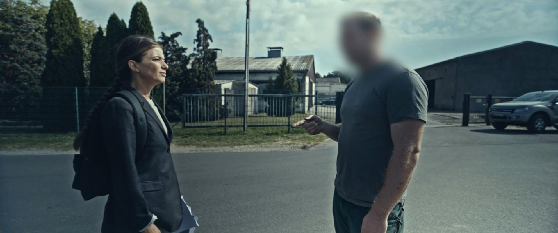 Eine Frau im Anzug spricht mit einem Mann vor Gebäuden und Autos.