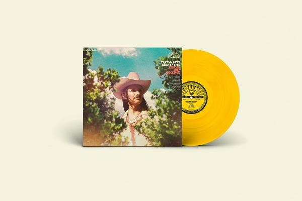Ein Mann mit Hut und langen Haaren blickt durch grüne Blätter auf ein gelbes Vinyl-Album.