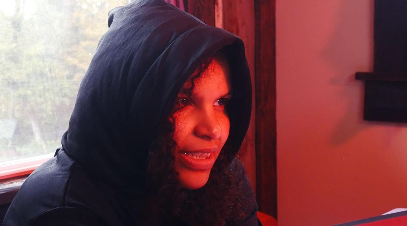 Eine Frau mit Kapuze blickt nach rechts, rot beleuchtet, vor einem Fenster.
