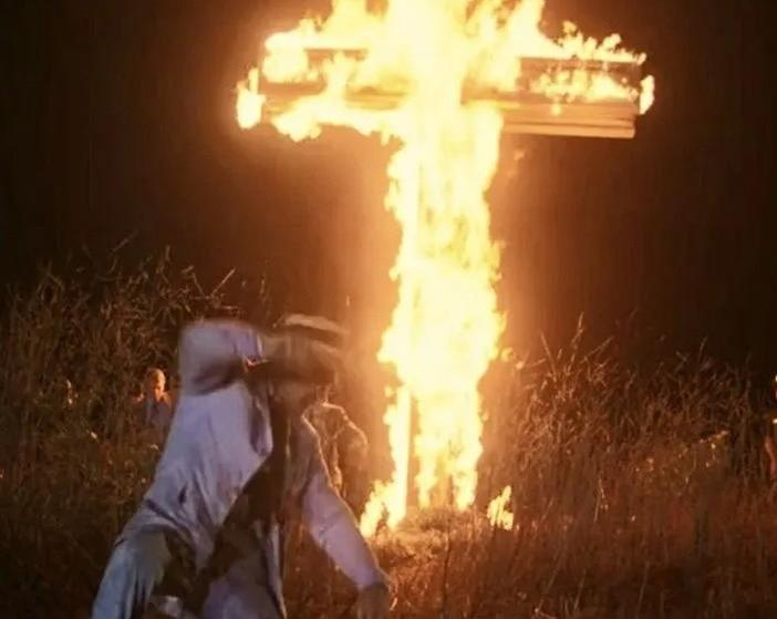 Ein Kreuz brennt nachts auf einem Feld, während eine Person davor steht.