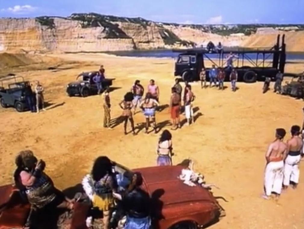 Menschen stehen um Autos und einen Lastwagen in einer sandigen Wüste mit Felsen im Hintergrund.