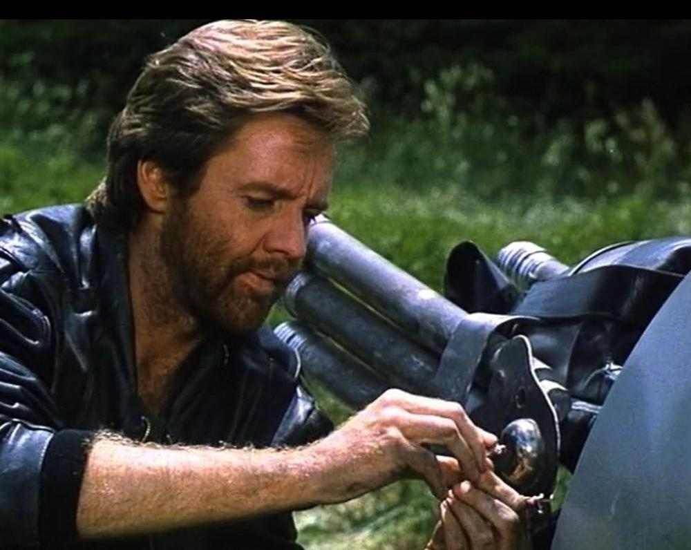 Ein Mann mit Bart in Lederjacke repariert eine Gatling-Kanone im Freien.