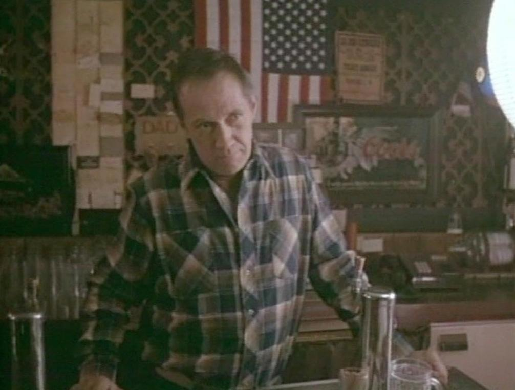 Ein Mann in kariertem Hemd steht hinter einer Bar mit einer US-Flagge.