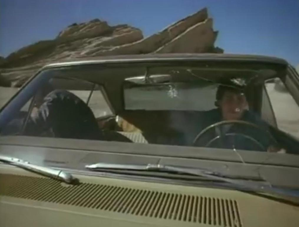Zwei Personen in einem Auto fahren durch eine Wüstenlandschaft mit Felsen.