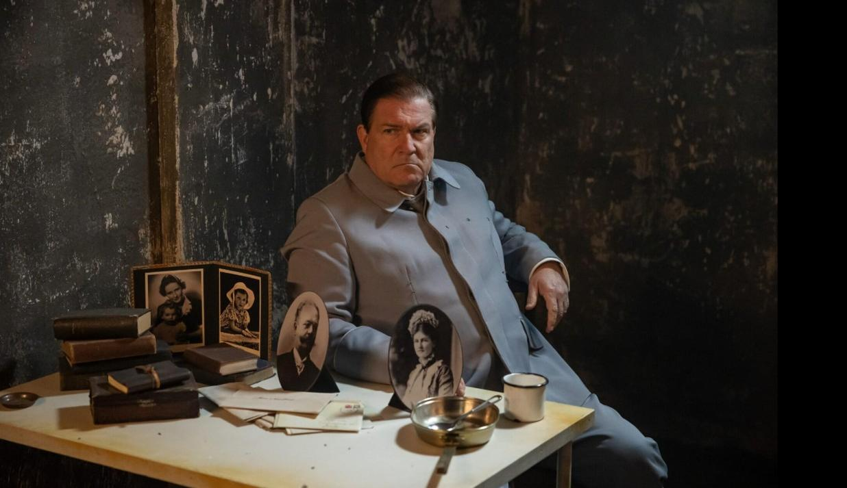 Ein Mann in einem grauen Mantel sitzt an einem Tisch mit alten Fotos und Büchern.