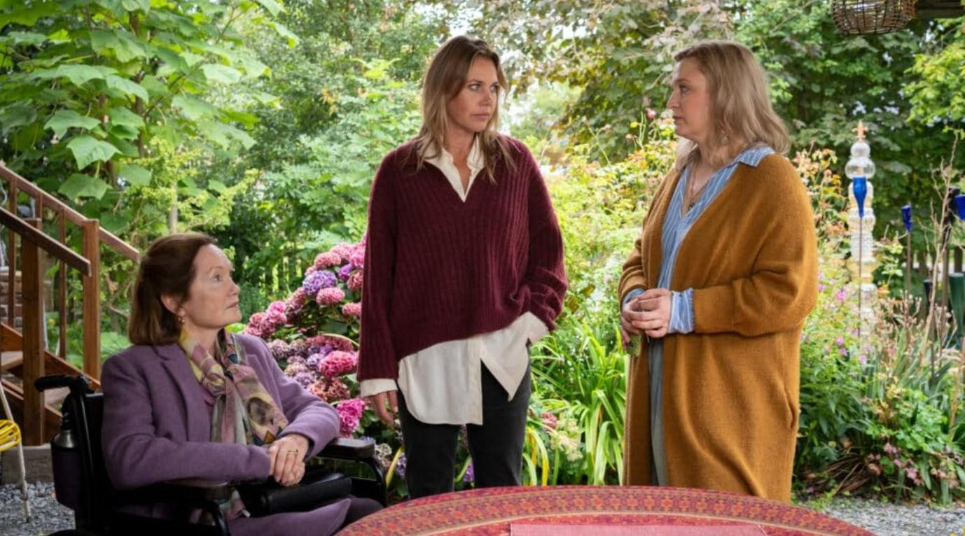 Drei Frauen stehen in einem üppigen Garten. Eine im Rollstuhl, eine im bordeauxroten Pullover, eine im ockergelben Cardigan.