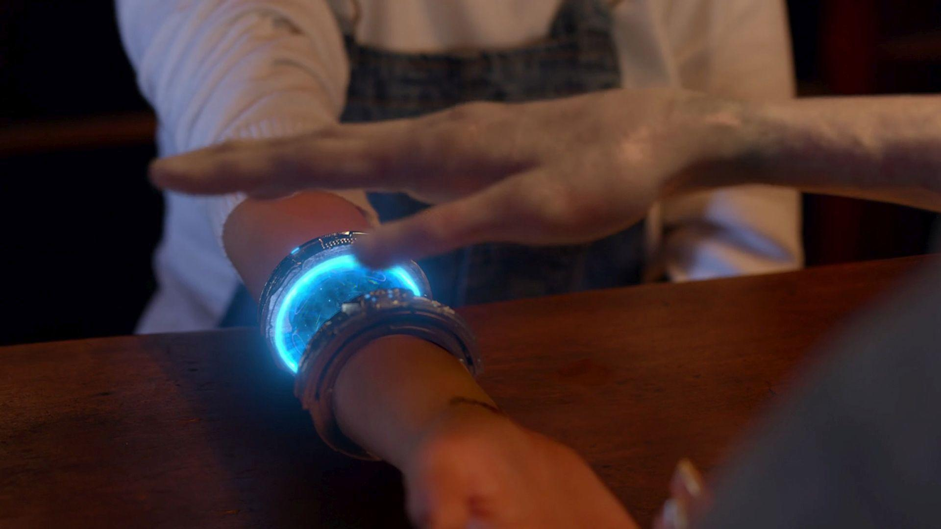 Ein leuchtendes blaues futuristisches Armband am Handgelenk, eine Hand schwebt darüber.