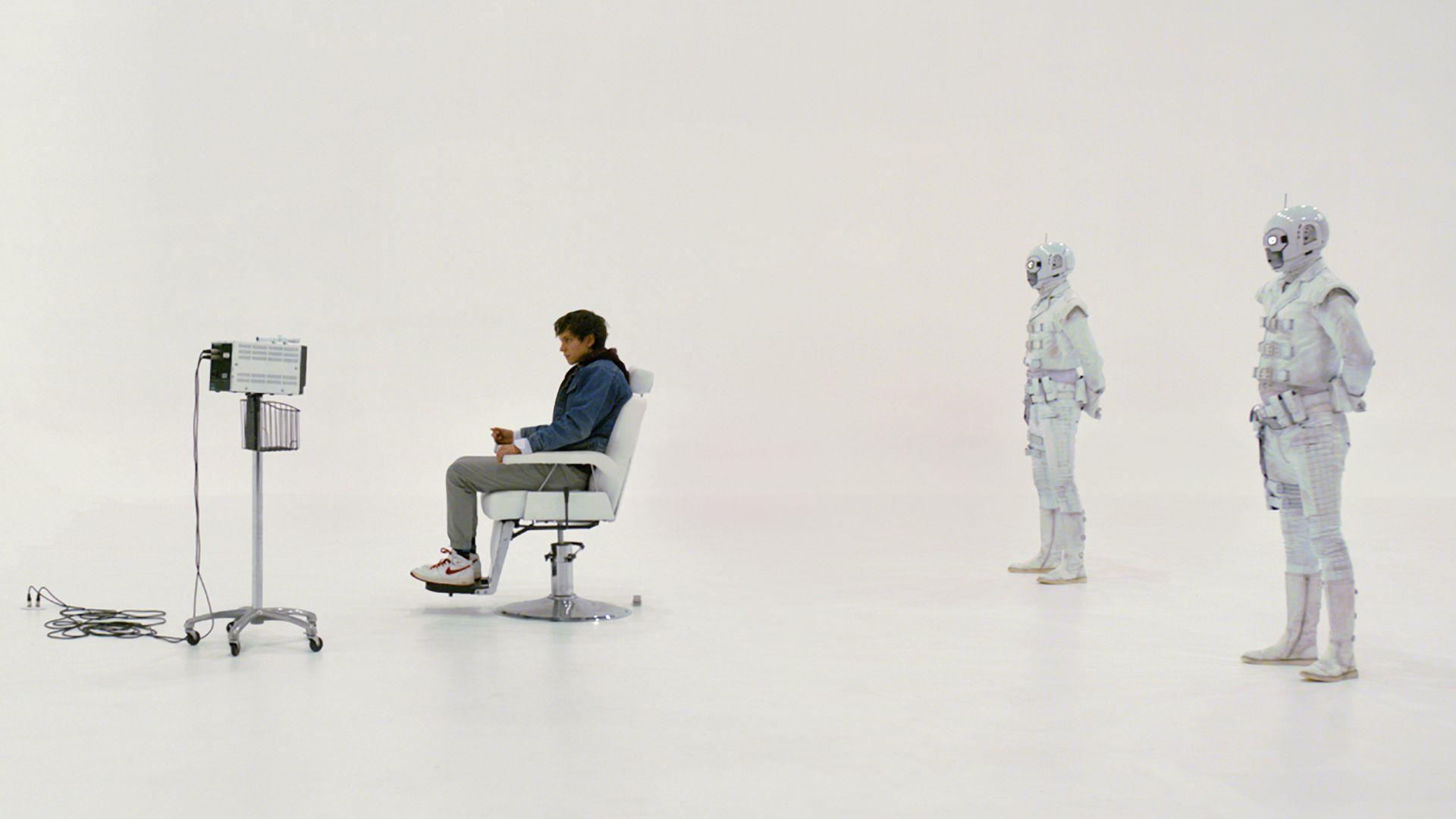 Ein Mann sitzt in einem Stuhl, zwei Roboter stehen dahinter im Weißen.